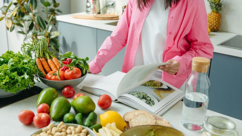 A person browsing a cookbook surrounded by fresh vegetables in a bright kitchen setting.